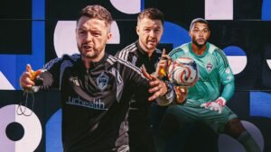 Matt Wells, newly appointed Colorado Rapids coach, stands confidently in training gear with players against a vibrant blue and black backdrop.