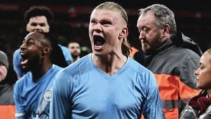 Erling Haaland celebrates passionately after scoring, wearing Manchester City's light blue kit, surrounded by teammates and staff.