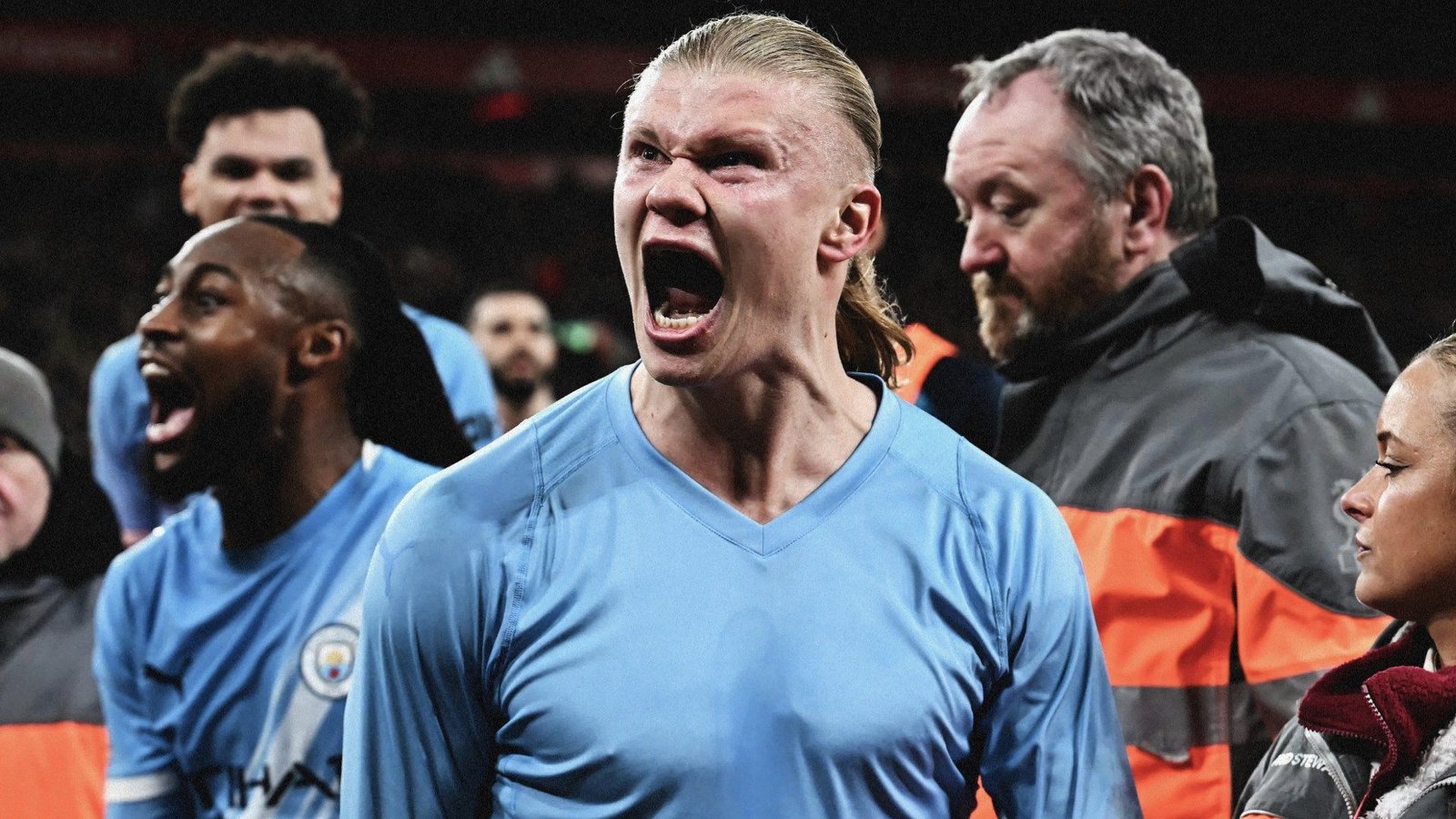 Erling Haaland celebrates passionately after scoring, wearing Manchester City's light blue kit, surrounded by teammates and staff.