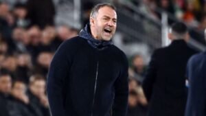 Hansi Flick, portant un pull sombre, gesticule avec passion en coachant en bord de terrain pendant un match, faisant preuve d'une concentration intense.