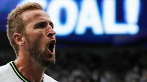 Harry Kane celebrates a goal with a passionate shout, wearing a Spurs kit, with "GOAL!" prominently displayed in the background.