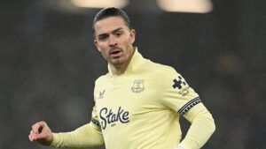 Jack Grealish en tenue extérieure d'Everton, l'air déterminé pendant un match, avec un fond de stade flou et des gouttes de pluie visibles.