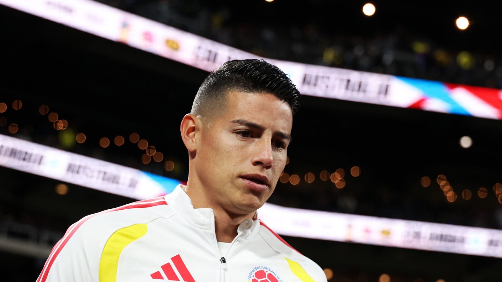 James Rodriguez en survêtement colombien, l'air pensif, entre sur le terrain, avec les projecteurs du stade et une foule enthousiaste en arrière-plan.