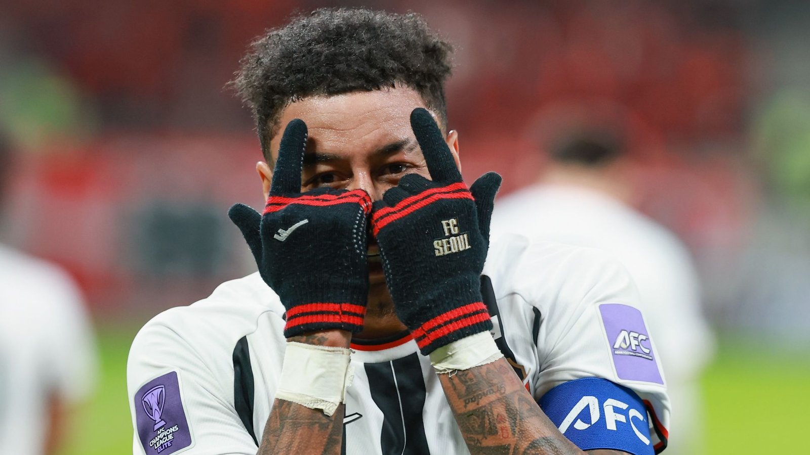 Jesse Lingard célèbre avec un geste espiègle, portant des gants du FC Séoul, dans un maillot d'équipe noir et blanc frappant pendant un match.