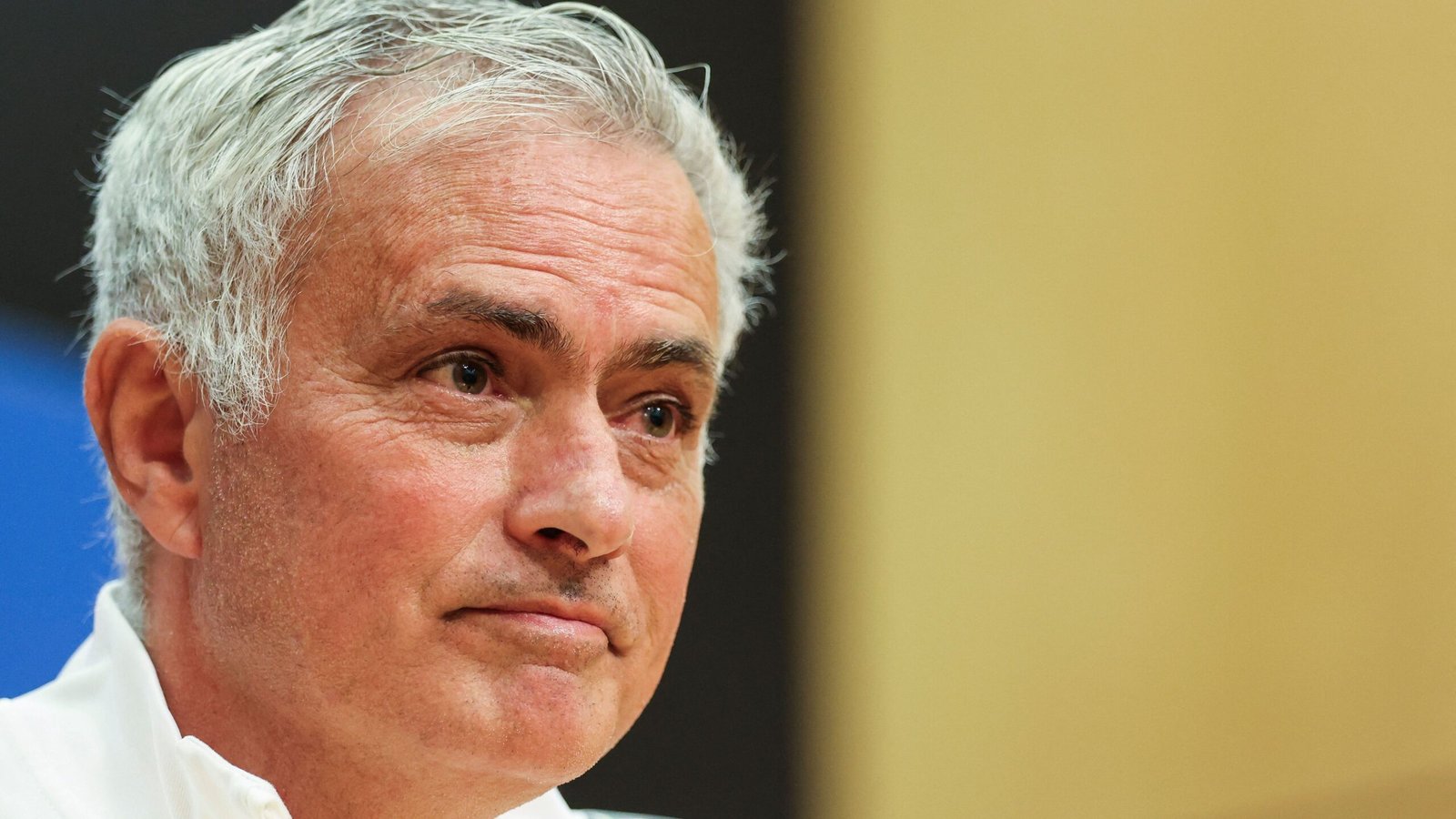 Jose Mourinho in a press conference, looking thoughtful, wearing a white jacket, with a neutral backdrop and soft lighting.