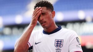 Jude Bellingham, in England's white kit with the Three Lions crest, looks distressed, holding his forehead during a match against Andorra.
