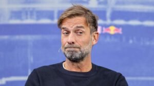 Jurgen Klopp appears thoughtful at a press conference, dressed in a black sweater, with a backdrop featuring Red Bull branding.