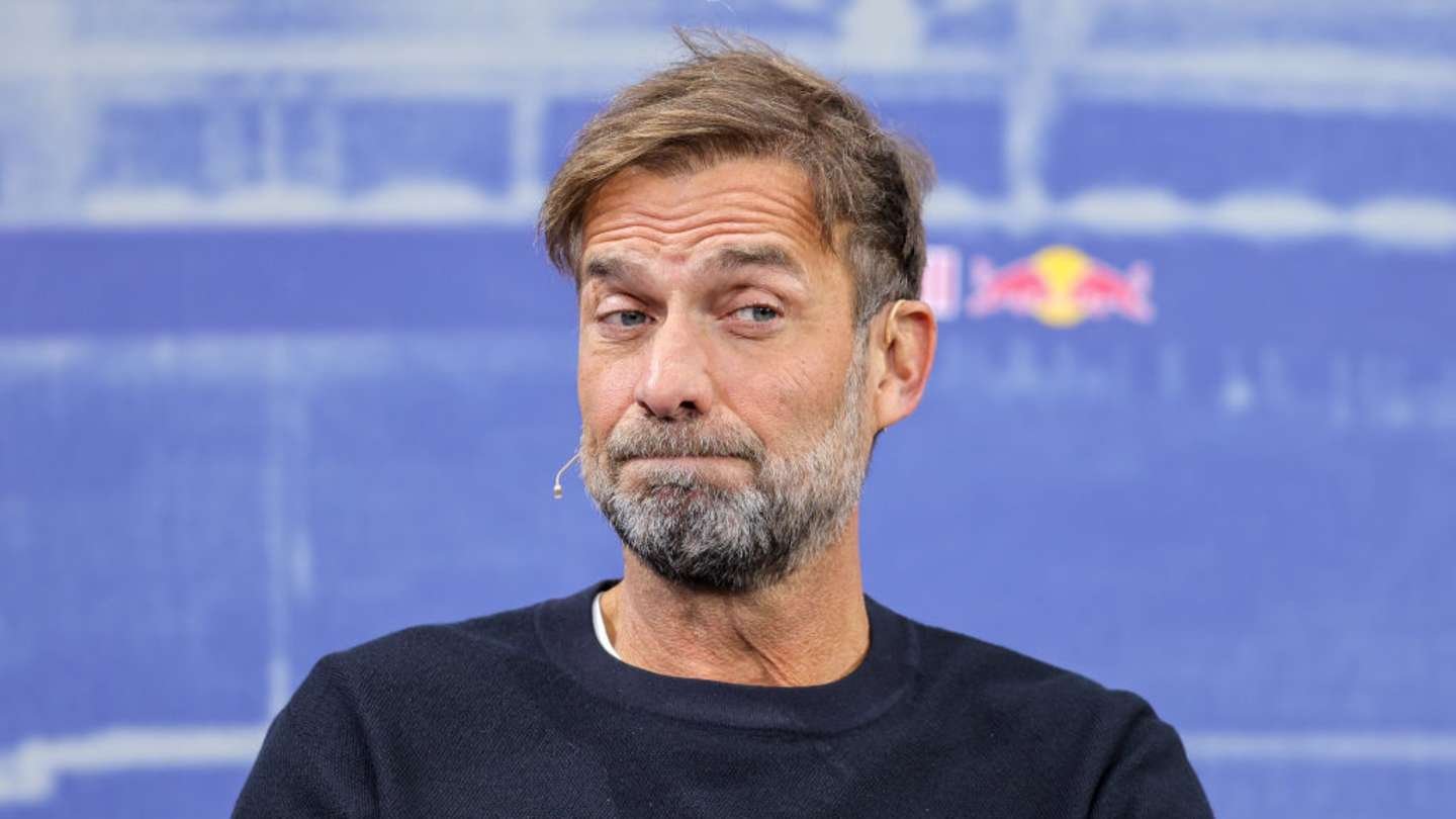 Jurgen Klopp appears thoughtful at a press conference, dressed in a black sweater, with a backdrop featuring Red Bull branding.