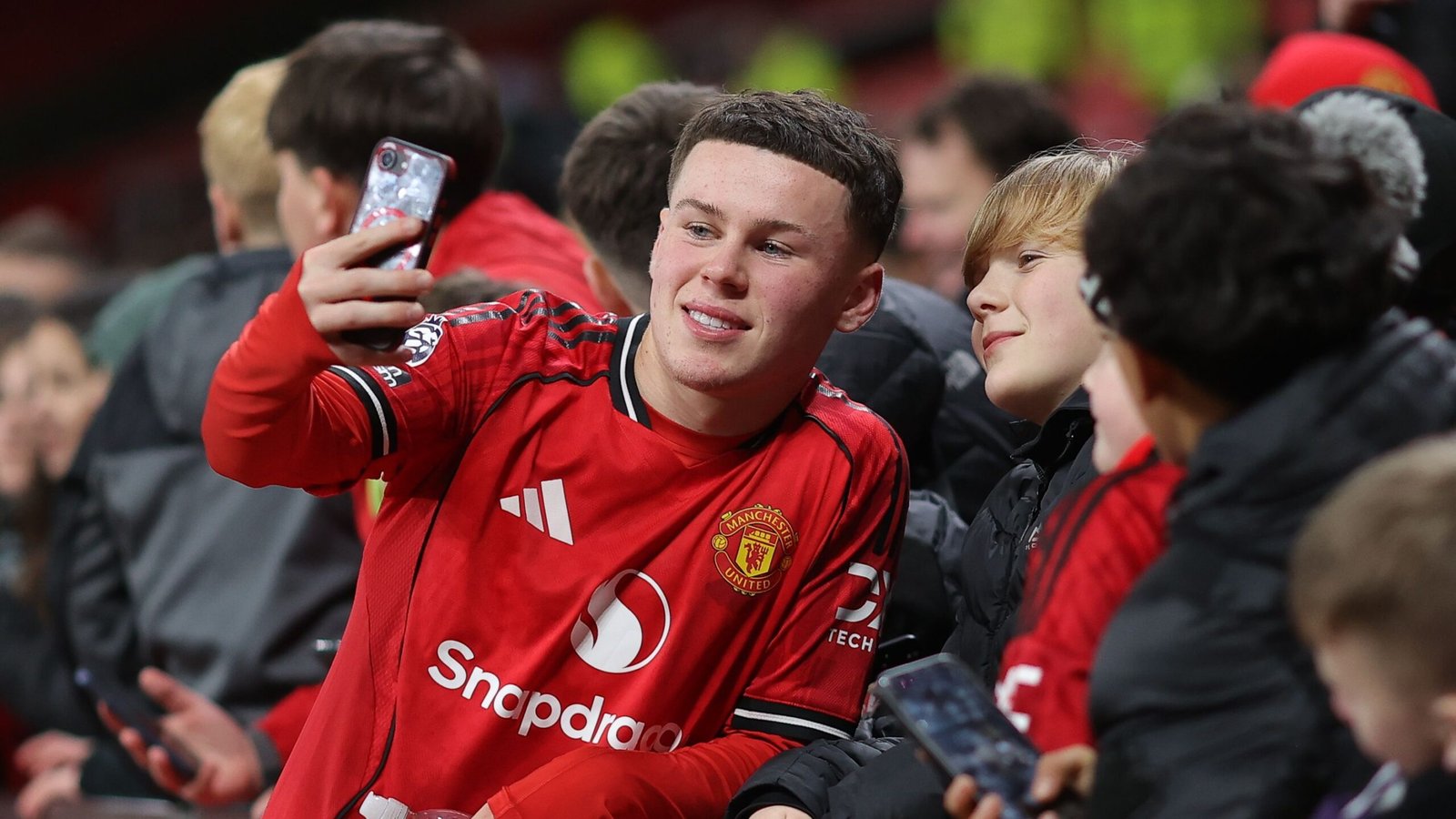 Kai Rooney maakt een selfie met opgewonden fans, allen gekleed in het rode tenue van Manchester United, en legt een moment van vreugde vast op Old Trafford.