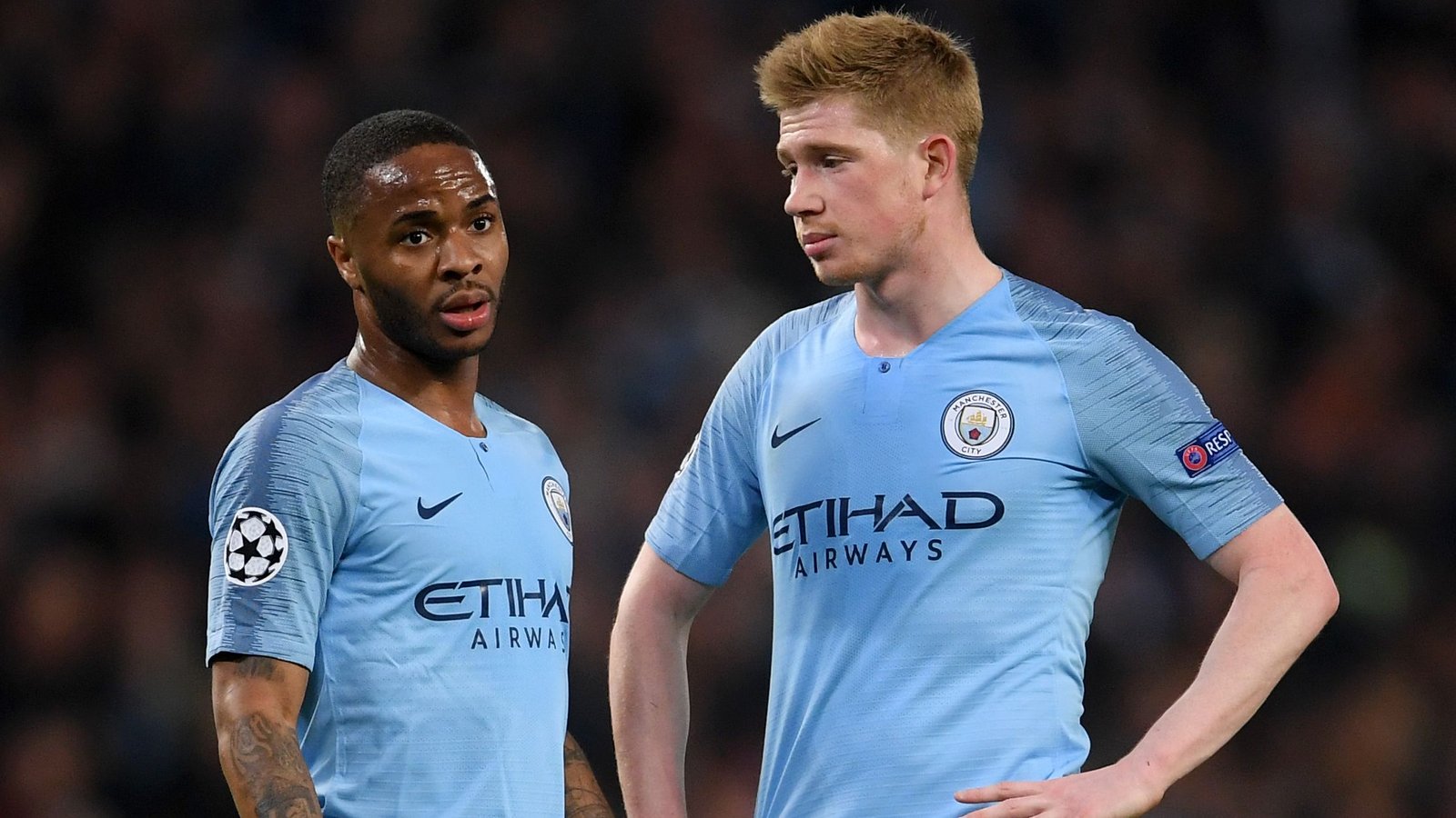 Raheem Sterling and Kevin De Bruyne in Manchester City's light blue kit during a Champions League match, looking focused on the pitch.