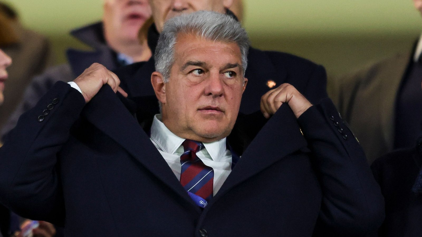 Joan Laporta ajustant sa veste dans les tribunes, l'expression concentrée au milieu de la foule, illustrant son rôle dans la direction du FC Barcelone.
