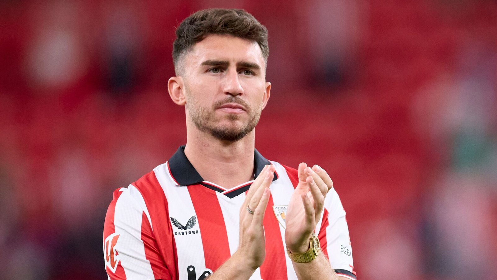 Aymeric Laporte applauding the crowd after a match, wearing a red and white striped jersey with a focused expression in a stadium setting.