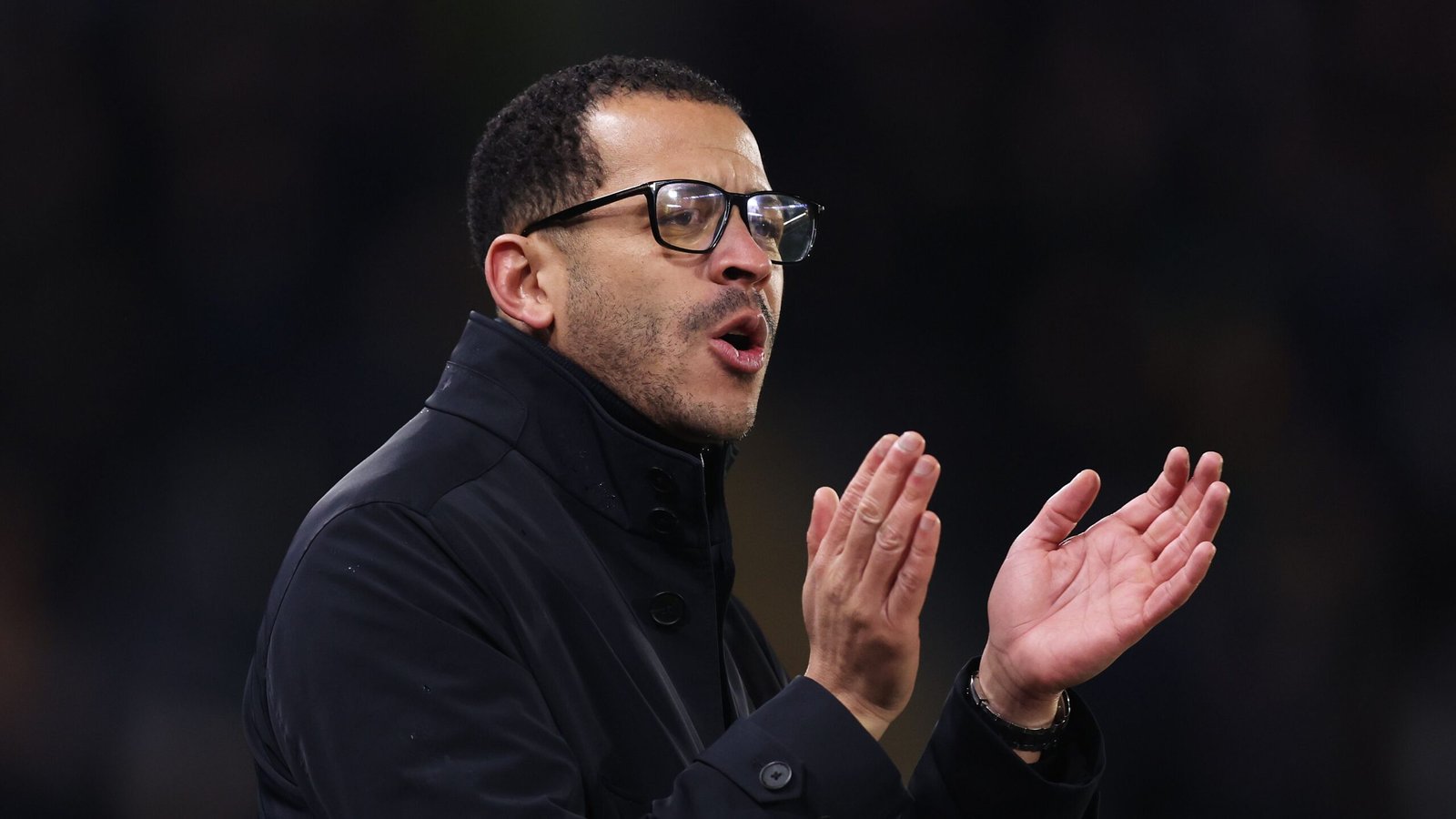 Liam Rosenior passionately gestures during a match, wearing glasses and a black coat, showing his commitment to his team.