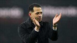 Liam Rosenior applauds the fans, wearing a black coat, in a stadium setting, showcasing his commitment to the team and Moroccan football.