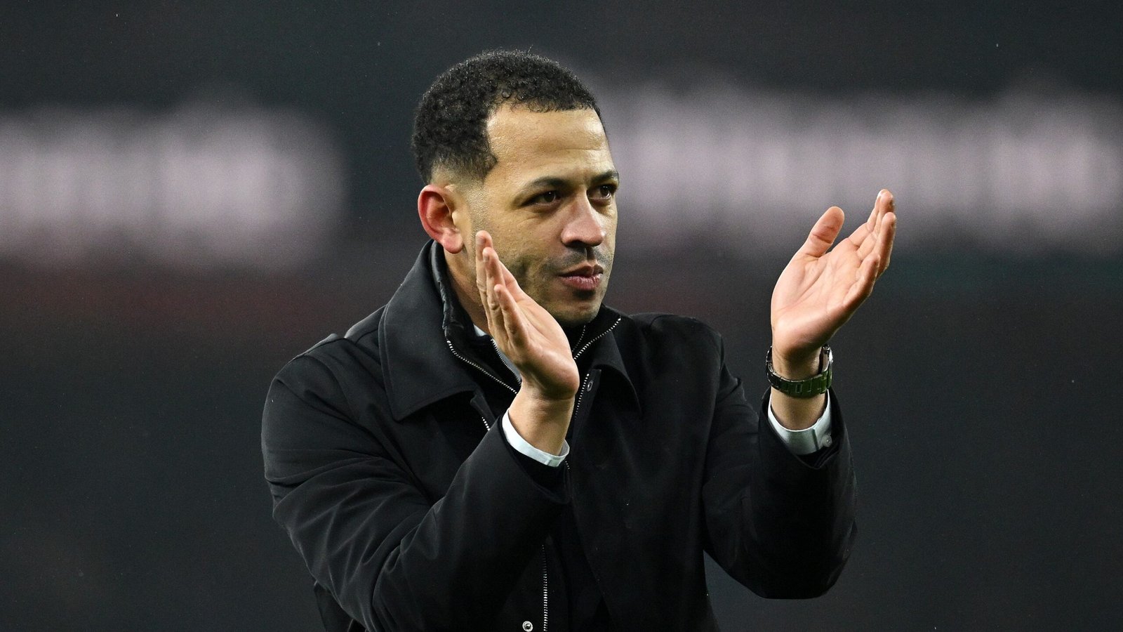 Liam Rosenior applauds the fans, wearing a black coat, in a stadium setting, showcasing his commitment to the team and Moroccan football.