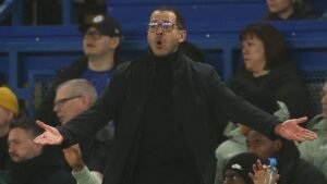 Liam Rosenior réagissant avec passion depuis la touche pendant un match, portant des lunettes et un manteau noir, avec des supporters visibles en arrière-plan.