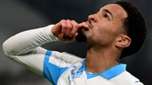 Le joueur de Marseille, Nwaneri, gesticule avec réflexion, portant le maillot blanc et bleu du club, soulignant un moment de concentration intense pendant un match.