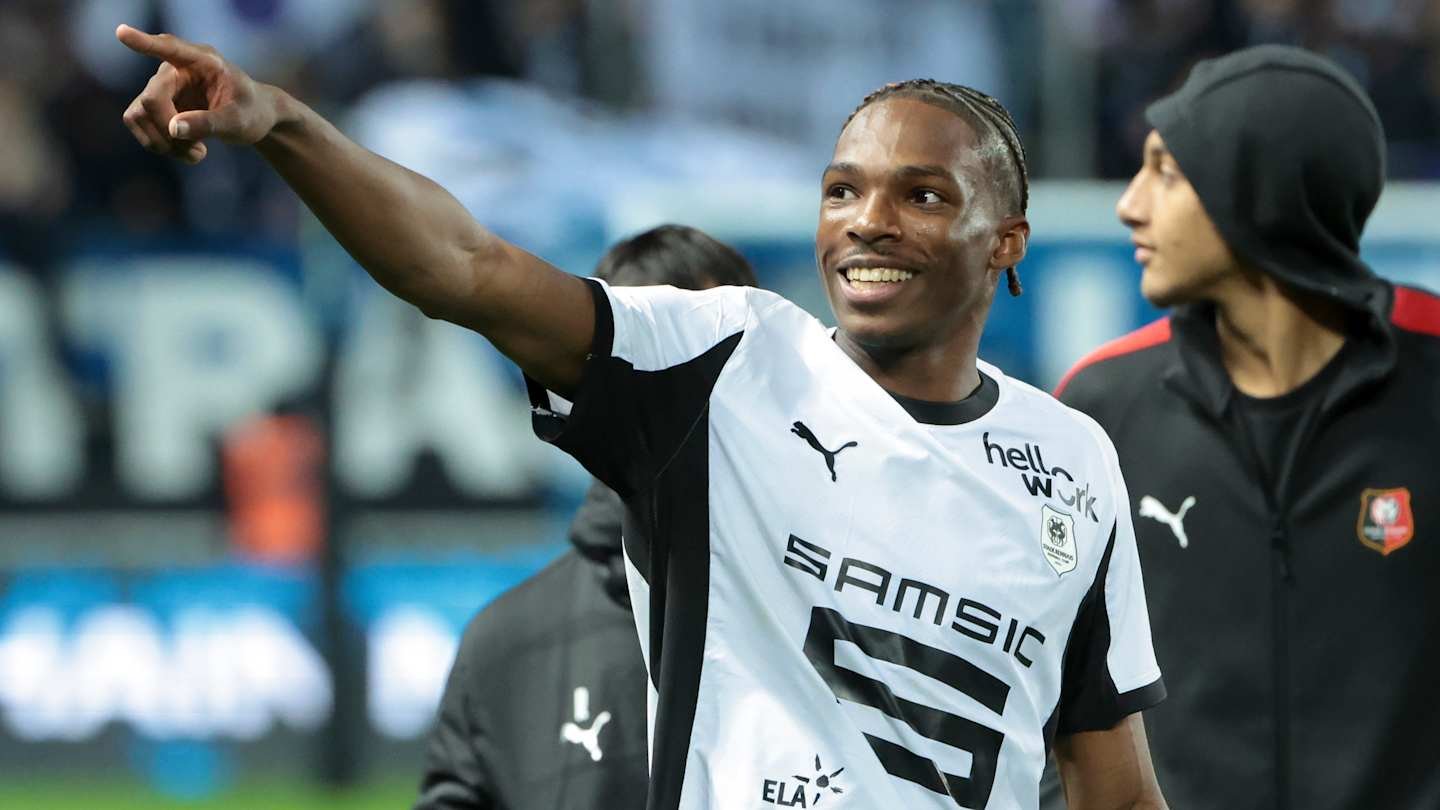 Rennes player smiling and pointing, wearing a black and white jersey, celebrating a moment after a match.