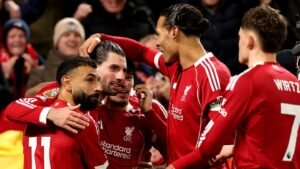 Liverpool players celebrate a goal, featuring Mohamed Salah, Virgil van Dijk, and Emre Can, in vibrant red kits amid cheering fans.