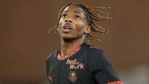 Jeremy Jacquet, gekleed in een zwart voetbaltenue met oranje accenten, kijkt geconcentreerd omhoog tijdens een wedstrijd voor zijn team.