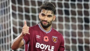 Lucas Paquetá celebrates in a maroon West Ham United kit, giving a thumbs up, with a football goal net in the background.