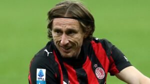 Luka Modric in AC Milan's black and red striped kit, smiling during a Serie A match. The background features a blurred stadium setting.