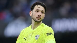Manchester City goalkeeper in a bright yellow kit, looking pensively during a match, highlighting the pressures of squad selection.
