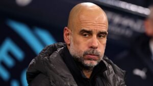 Pep Guardiola apparaît pensif sur le bord du terrain, l'air concentré et vêtu d'une veste grise, se détachant sur un fond de stade flou.
