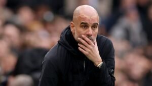 Pep Guardiola in overpeinzingen tijdens een wedstrijd, gekleed in een zwart jasje, met een vervaagde menigte op de achtergrond in een voetbalstadion.