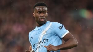 Marc Guehi dans le maillot bleu clair de Manchester City, l'air perplexe sur le terrain pendant un match, avec un arrière-plan flou de supporters.