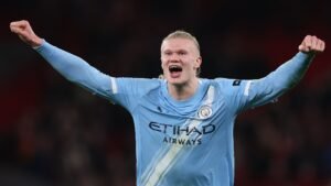 Erling Haaland célébrant avec passion dans le maillot bleu clair de Manchester City après un but, exhibant ses cheveux longs distinctifs et son exaltation.