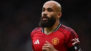 Joueur de Manchester United au crâne rasé et portant une barbe, vêtu du maillot rouge de l'équipe avec une bande noire, concentré pendant un match.