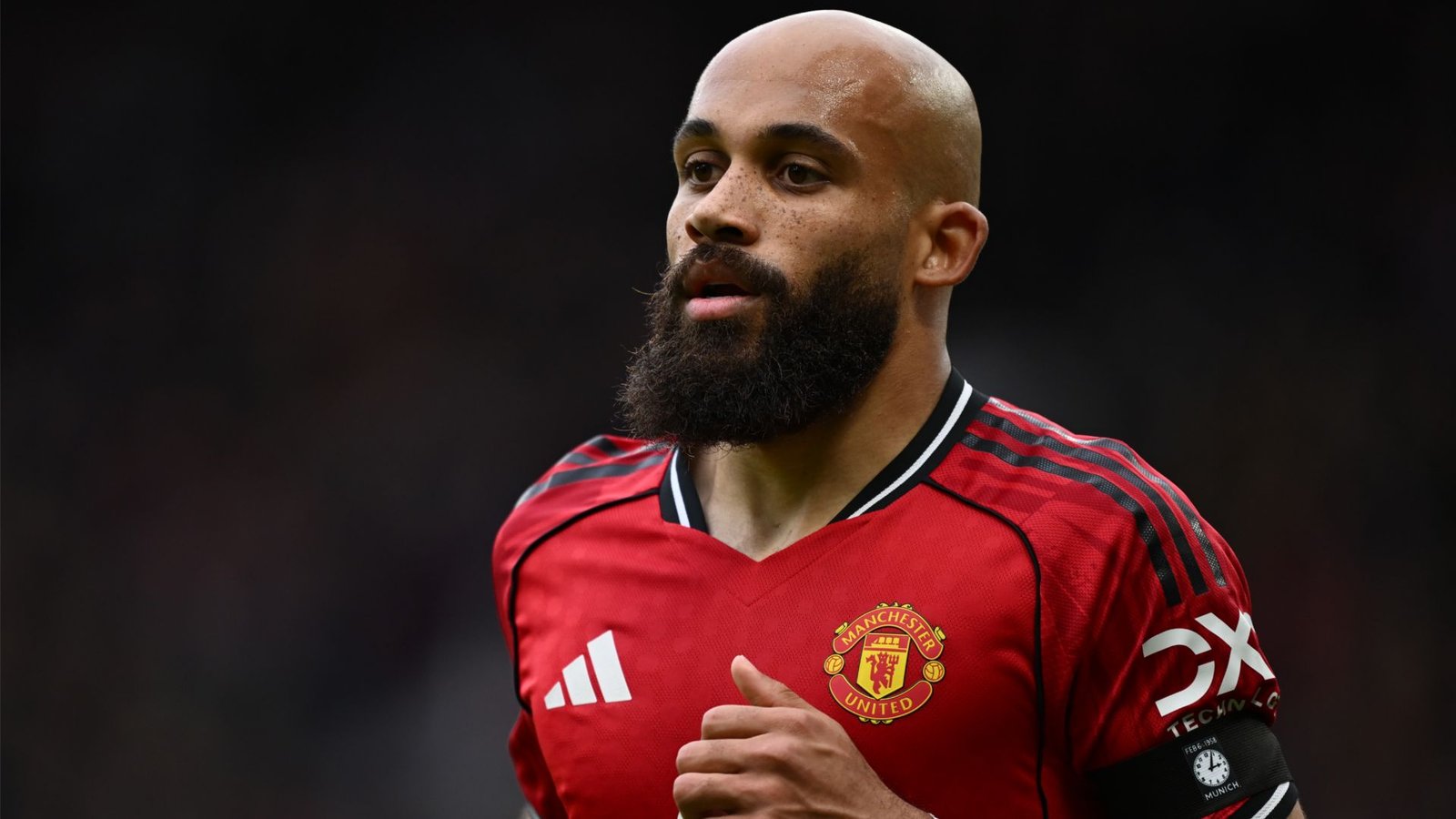 Joueur de Manchester United au crâne rasé et portant une barbe, vêtu du maillot rouge de l'équipe avec une bande noire, concentré pendant un match.