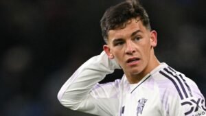 Shea Lacey, vêtu du maillot blanc de Manchester United, semble pensif pendant un match, faisant preuve de détermination et de concentration.