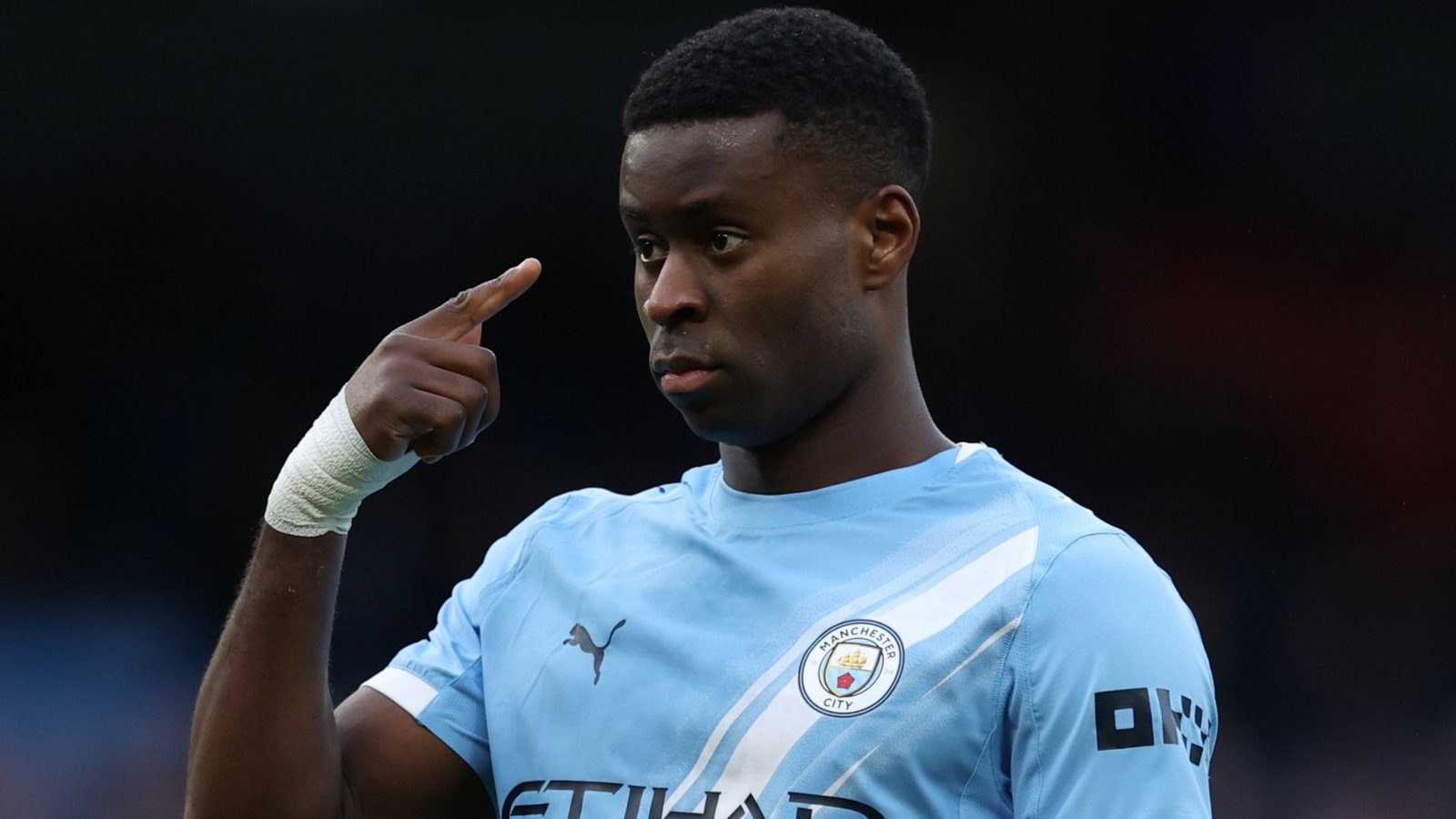 Marc Guehi esquisse un geste avec une expression déterminée, portant le maillot bleu clair de Manchester City pendant un match, reflétant sa concentration et son engagement.