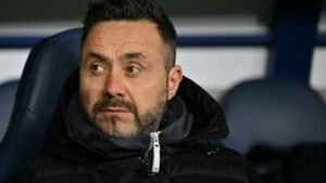 Roberto De Zerbi, gekleed in een zwarte jas, kijkt bedachtzaam vanaf de bank tijdens een wedstrijd.