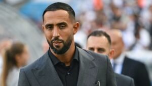 Medhi Benatia in a smart grey suit, looking serious as he arrives at a football match, showcasing his role in Moroccan football.