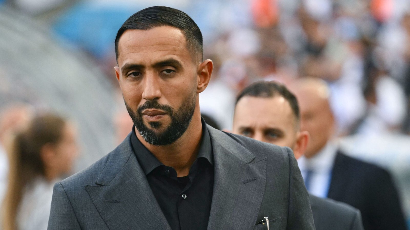 Medhi Benatia in a smart grey suit, looking serious as he arrives at a football match, showcasing his role in Moroccan football.