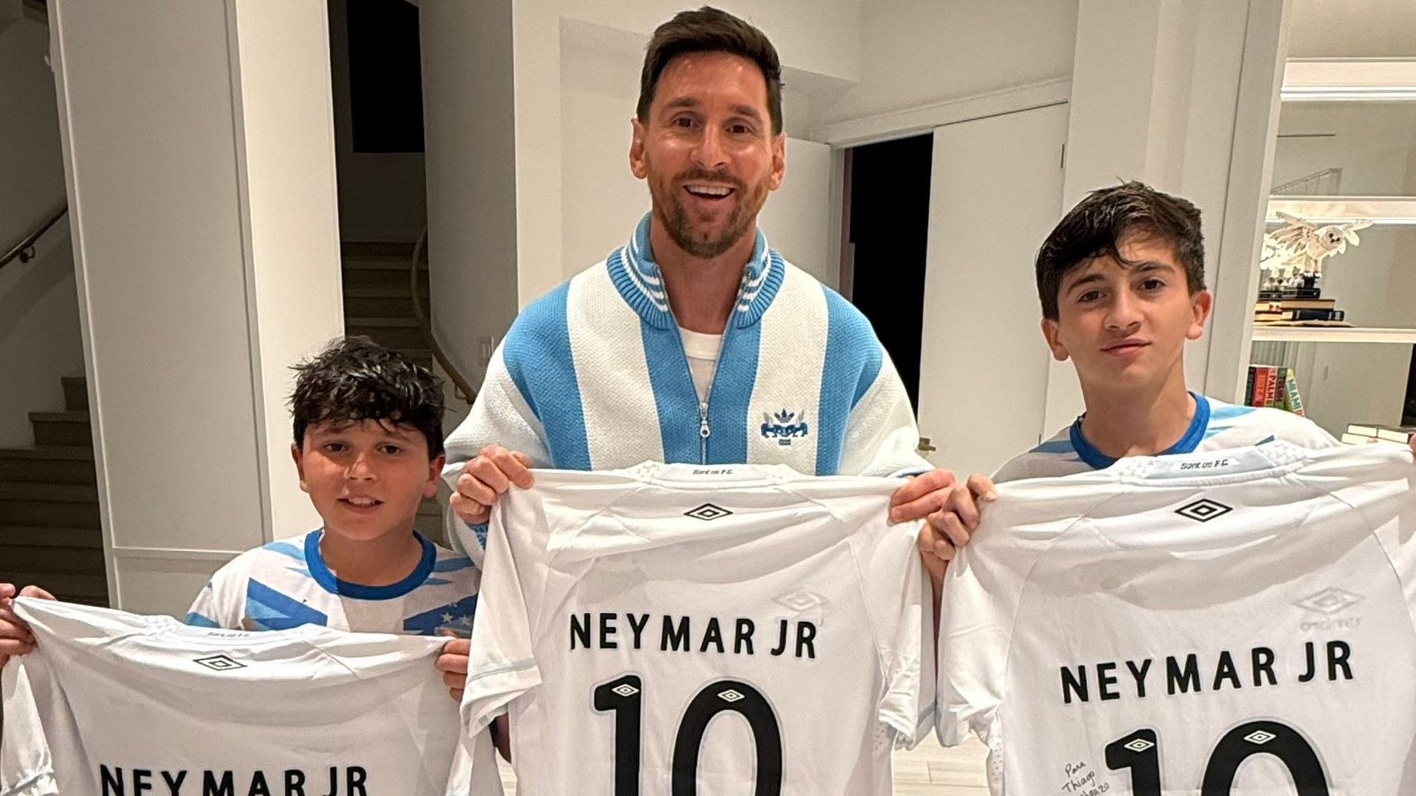 Messi holds up Neymar Jr. shirts with his two sons, all wearing Argentina kits, in a bright indoor setting.