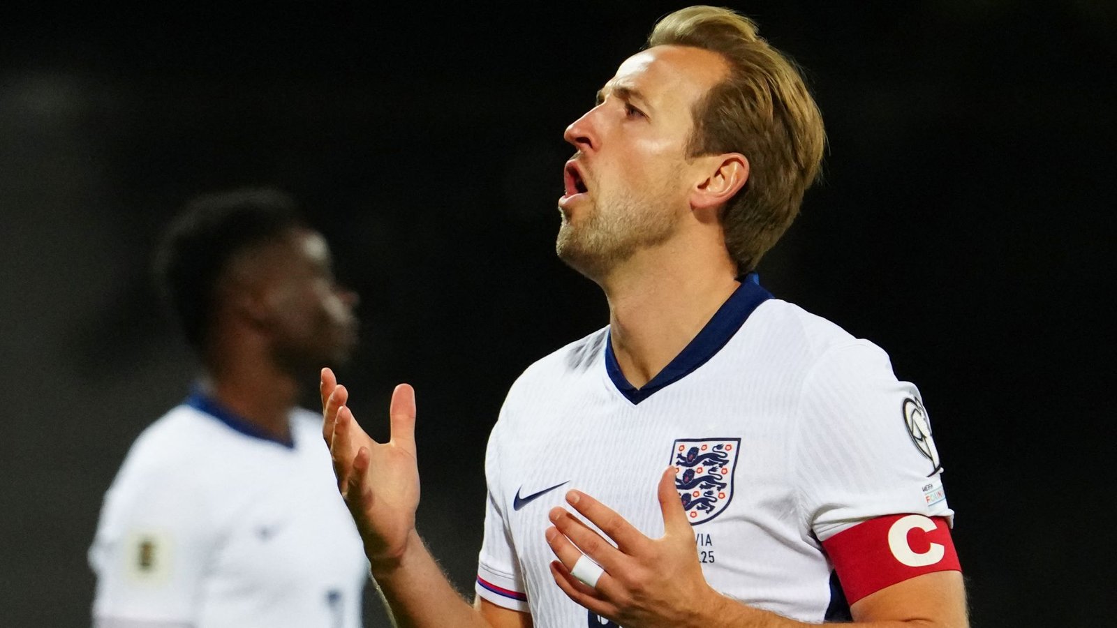Harry Kane, wearing the England captain's armband, shows frustration during a match, with a teammate in the background.