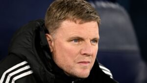 Eddie Howe, entraîneur de Newcastle United, concentré et déterminé, portant une veste noire à rayures blanches, assis sur le banc.