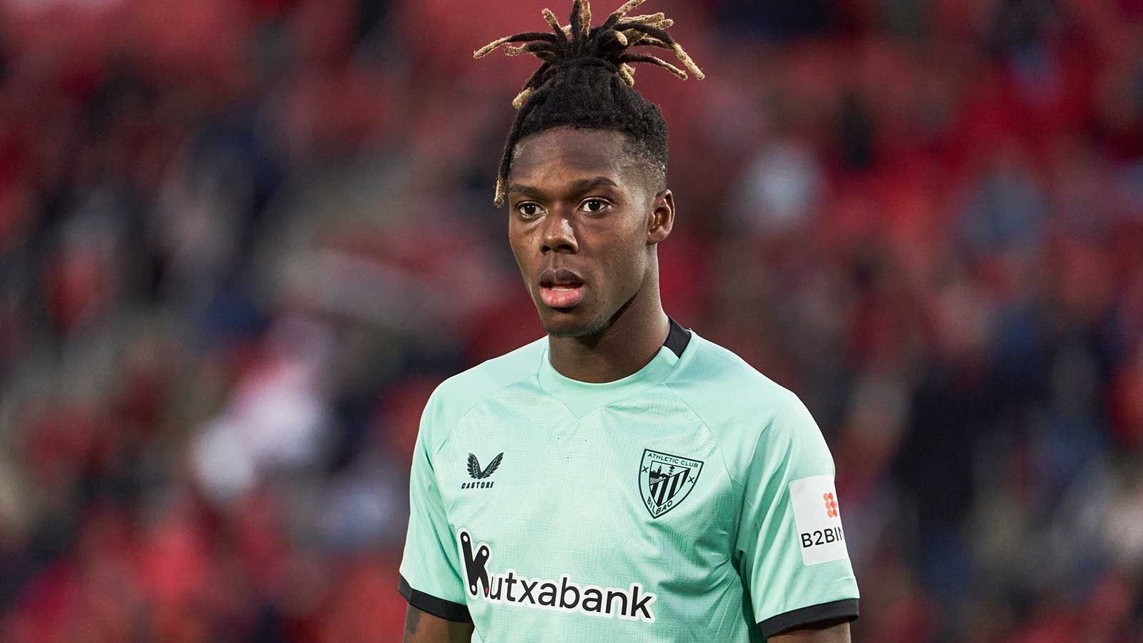 Nico Williams portant un maillot de l'Athletic Bilbao vert menthe, l'air inquiet lors d'un match, devant des sièges de stade rouges.