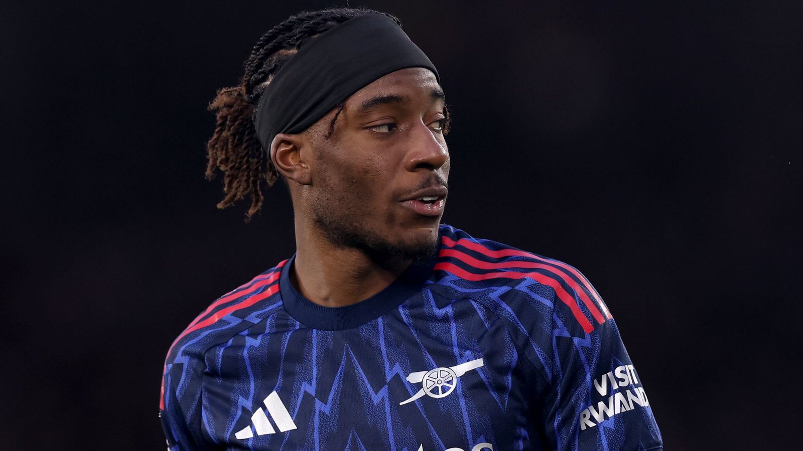 Noni Madueke, vêtu d'un maillot bleu et rouge, affichant une détermination sur le terrain pendant un match, démontrant sa concentration et son talent footballistique.