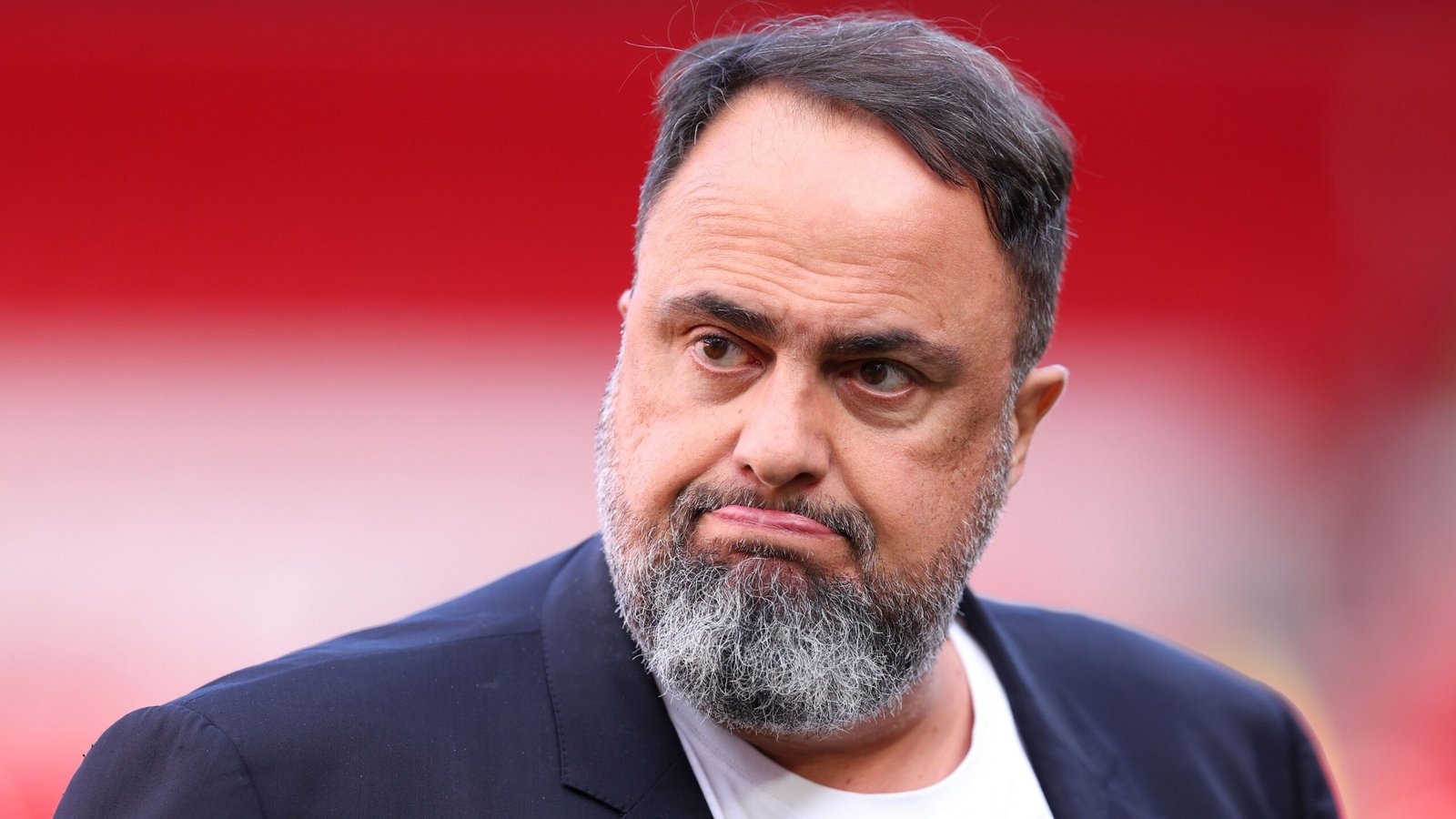 De toekomstige manager van Nottingham Forest, met een serieuze uitdrukking en een baard, staat tegen een wazige rode stadionachtergrond.