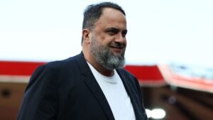Marinakis sourit avec assurance, vêtu d'une élégante veste noire sur une chemise blanche, devant un fond de stade.