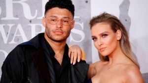 Alex Oxlade-Chamberlain pose avec Perrie Edwards sur un tapis rouge, tous deux vêtus de tenues élégantes avec un fond raffiné.