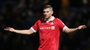 L'attaquant de Wrexham, Paul Mullin, vêtu du maillot domicile rouge, les bras grands ouverts, exprimant sa frustration lors d'un match.