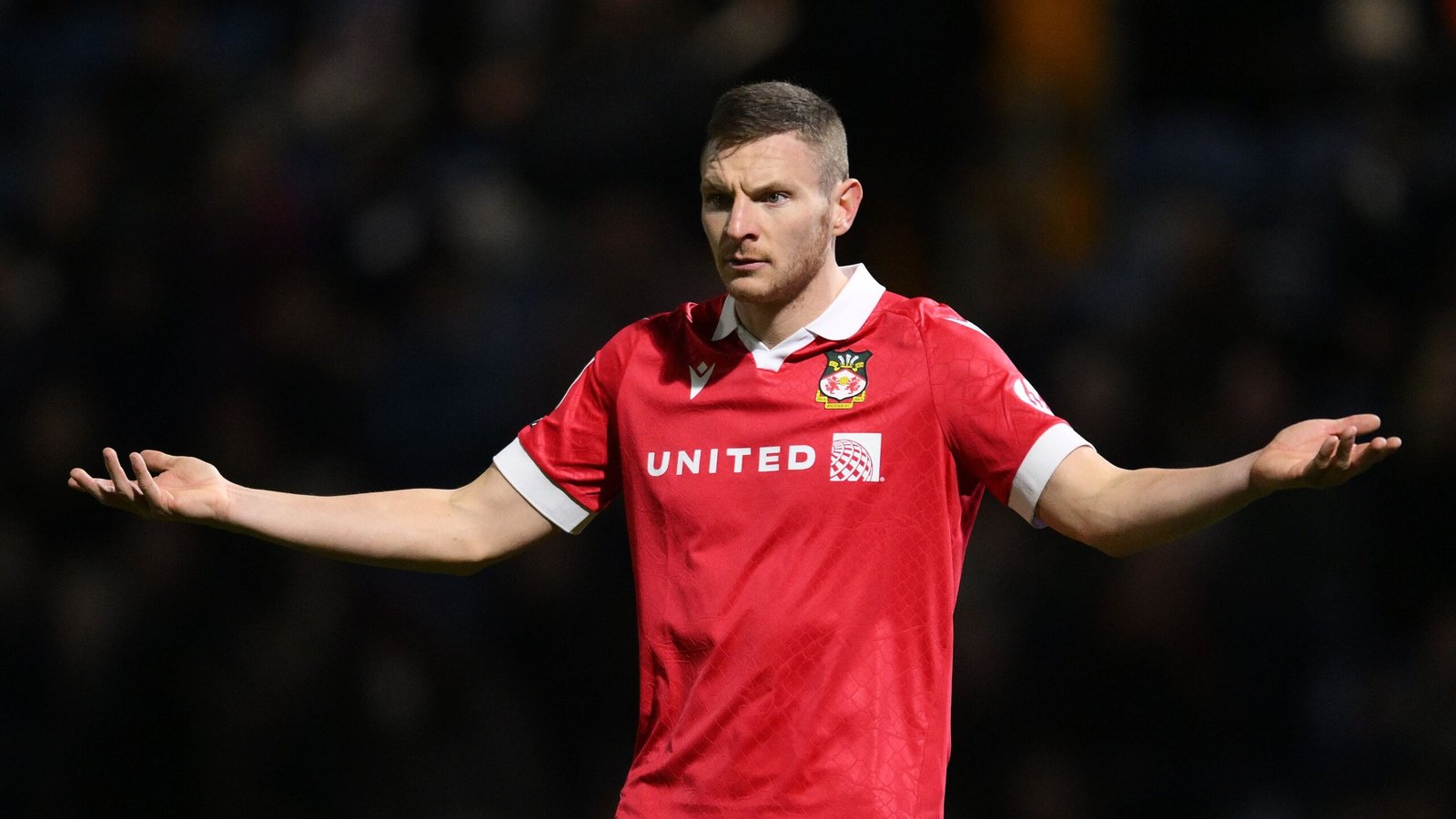 L'attaquant de Wrexham, Paul Mullin, vêtu du maillot domicile rouge, les bras grands ouverts, exprimant sa frustration lors d'un match.