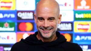 Pep Guardiola smiling during a press conference, with a colourful backdrop featuring Champions League logos and sponsorships.