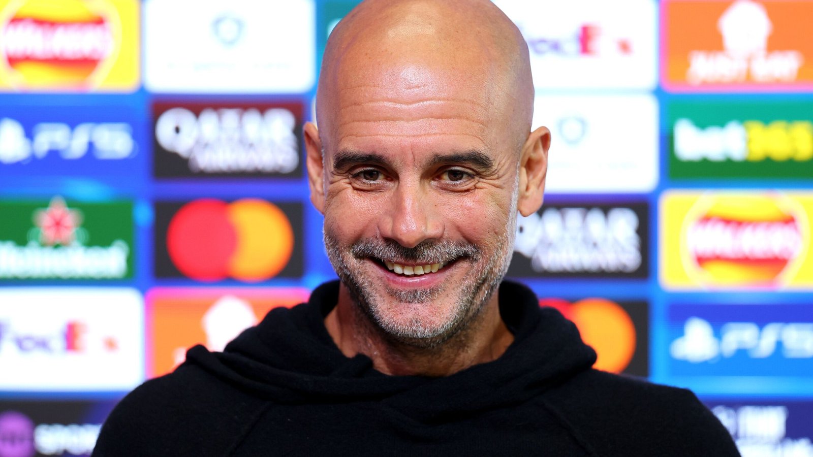 Pep Guardiola smiling during a press conference, with a colourful backdrop featuring Champions League logos and sponsorships.